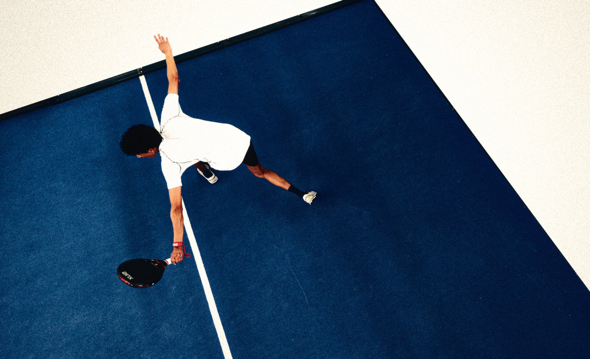Padel player stretching for a shot in the corner of a blue court, seen from above