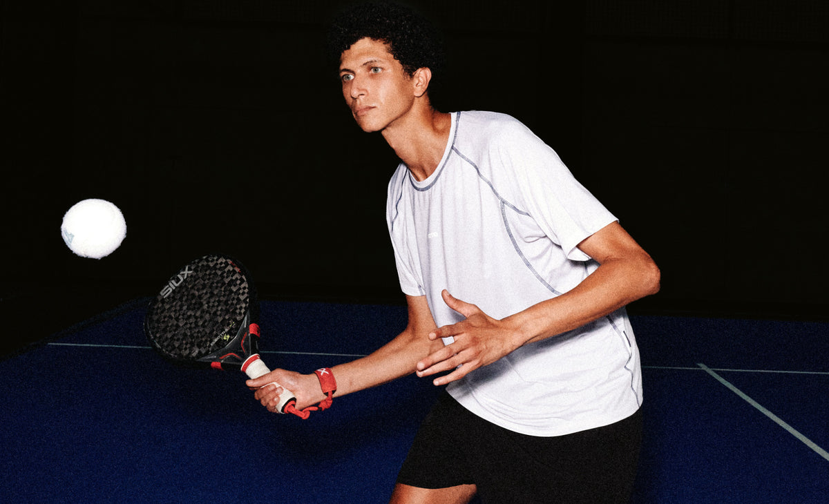 Padel player in the white TIOTIO Performance Tee returning a ball on a blue indoor court