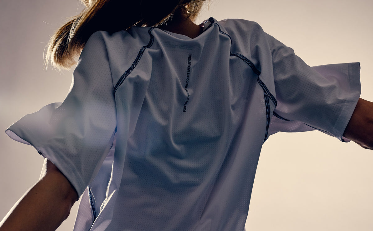 Athlete from behind in the white Performance Tee, shoulder print visible against a pale studio wall